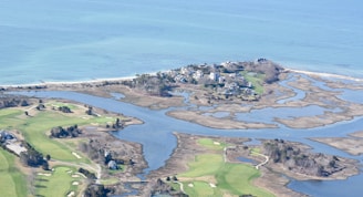 An aerial view of a coastal area featuring a small peninsula with houses nestled among trees. Surrounding the peninsula are winding waterways and marshland. Adjacent to this is a golf course with neatly cut grass and sand pits. The ocean in the background is a calm blue, adding a serene backdrop to the landscape.