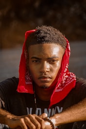 A person is sitting and looking directly at the camera, wearing a red bandana wrapped around their head. The background is obscured, putting emphasis on the focused expression and details of the face.