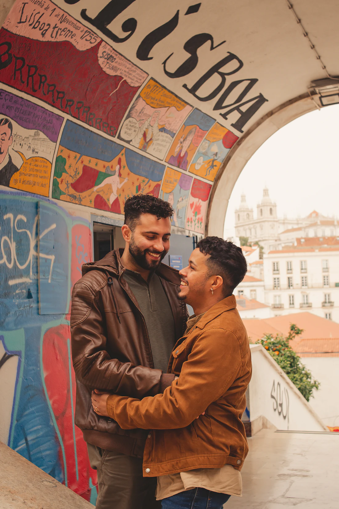 Julian and Mateo laughing in an embrace under a painted archway above the rooftops of Lisbon