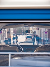 Inside view of a vintage vehicle with sheepskin seat covers, looking through the windshield at a bustling city street. People and cyclists pass by on the road, alongside parked cars. The scene suggests a lively urban environment.