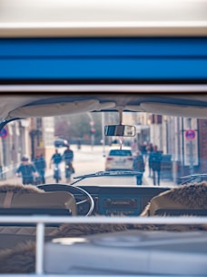 Inside view of a vintage vehicle with sheepskin seat covers, looking through the windshield at a bustling city street. People and cyclists pass by on the road, alongside parked cars. The scene suggests a lively urban environment.