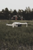 A white drone is flying over a grassy field with blurred trees in the background. The focus is on the drone, highlighting its sleek design and propellers in motion.