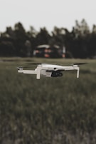 A white drone is flying over a grassy field with blurred trees in the background. The focus is on the drone, highlighting its sleek design and propellers in motion.