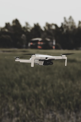 A white drone is flying over a grassy field with blurred trees in the background. The focus is on the drone, highlighting its sleek design and propellers in motion.