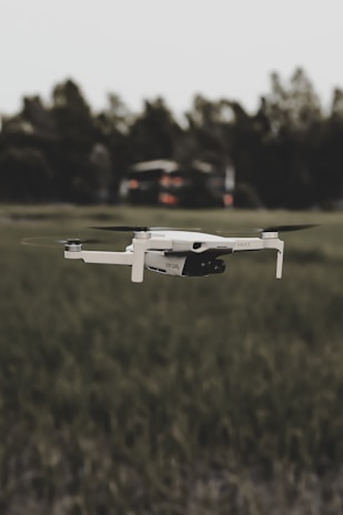 A white drone is flying over a grassy field with blurred trees in the background. The focus is on the drone, highlighting its sleek design and propellers in motion.