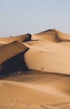 A vast desert landscape with rolling sand dunes. A person riding an off-road vehicle creates tracks across the dunes, capturing a sense of adventure and solitude.