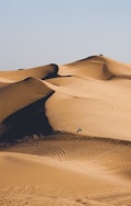 A vast desert landscape with rolling sand dunes. A person riding an off-road vehicle creates tracks across the dunes, capturing a sense of adventure and solitude.