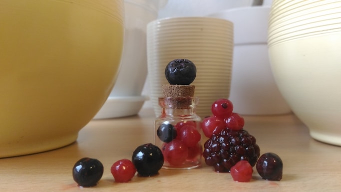 A small glass jar stands on a wooden surface with a cork lid, surrounded by a selection of berries including blackberries, blueberries, and red currants. In the background, several ceramic bowls in cream and white tones are visible, creating a simple and clean setting.