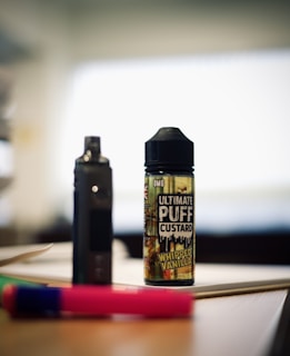 A bottle labeled 'Ultimate Puff Custard Whipped Vanilla' is placed on a table alongside a dark device that resembles a vape. The background is blurred, creating a focused view of the bottle and device. Additionally, a pink and green pen lies on the table nearby.