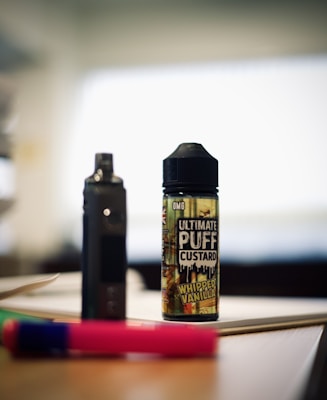 A bottle labeled 'Ultimate Puff Custard Whipped Vanilla' is placed on a table alongside a dark device that resembles a vape. The background is blurred, creating a focused view of the bottle and device. Additionally, a pink and green pen lies on the table nearby.