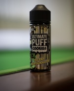 A bottle labeled 'Ultimate Puff Cookies' with the flavor 'Creamy Marshmallow' printed. It stands on a flat surface, with a blurred green and brown background suggesting a relaxed setting.