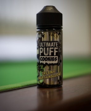A bottle labeled 'Ultimate Puff Cookies' with the flavor 'Creamy Marshmallow' printed. It stands on a flat surface, with a blurred green and brown background suggesting a relaxed setting.