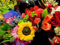 A vibrant and colorful assortment of flower bouquets with roses, sunflowers, and various green leaves. The arrangement includes red, orange, and pink roses, complemented by green foliage and small decorative berries. Bright yellow sunflowers and purple thistles add contrast and variety to the collection.