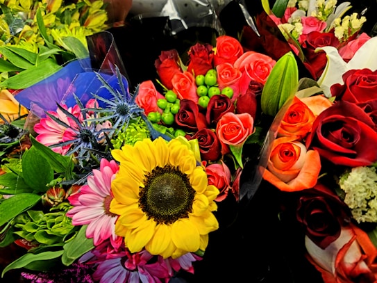 A vibrant and colorful assortment of flower bouquets with roses, sunflowers, and various green leaves. The arrangement includes red, orange, and pink roses, complemented by green foliage and small decorative berries. Bright yellow sunflowers and purple thistles add contrast and variety to the collection.