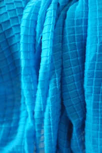 A close-up view of a textured fabric with a grid pattern. The fabric appears to be draped or folded, with visible shadows and highlights accentuating the three-dimensional quality of the material.