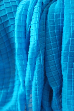 A close-up view of a textured fabric with a grid pattern. The fabric appears to be draped or folded, with visible shadows and highlights accentuating the three-dimensional quality of the material.