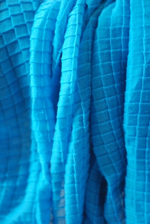 A close-up view of a textured fabric with a grid pattern. The fabric appears to be draped or folded, with visible shadows and highlights accentuating the three-dimensional quality of the material.