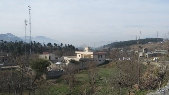 A rural landscape featuring a small settlement with scattered houses and buildings, surrounded by fields and trees. A tall communication tower is visible on the left side, and distant mountains can be seen in the background under a slightly overcast sky.