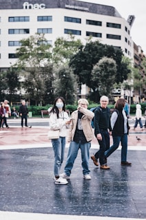 A group of people walking in an urban area, with two individuals at the forefront appearing engaged with a phone. They are surrounded by modern architecture, including a building with trees lining the sidewalk. The atmosphere appears casual and lively.