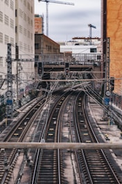 A multi-track railway passes through an urban area with tall buildings on either side. Overhead wires and signal lights are visible, along with graffiti-covered walls. In the background, construction cranes and a bridge are present.