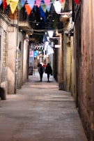 A narrow, cobblestone alleyway is adorned with colorful triangular flags hanging overhead. Two figures are walking down the alley, illuminated by the soft glow of street lamps and strings of small lights. The buildings lining the alley appear old and rustic, with some signs and balconies visible.