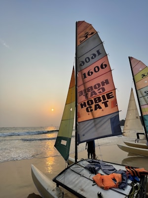 Catamarans with colorful sails are positioned on a sandy beach as the sun sets over the ocean. The sails display various designs and numbers, and the water gently meets the shoreline. Life jackets and other equipment are visible on one of the catamarans.
