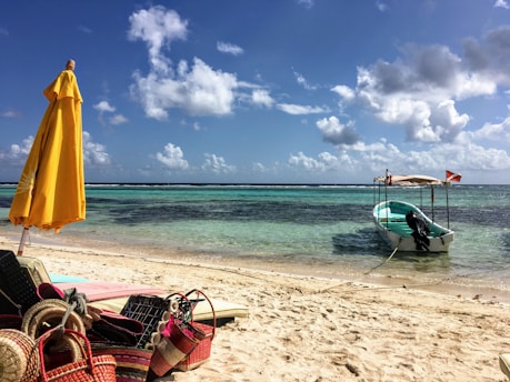 A sunny beach scene with a yellow umbrella on the sand and various colorful beach items like bags and mats nearby. A small boat tied by a rope is floating on clear turquoise water. The sky is bright blue with scattered fluffy white clouds.