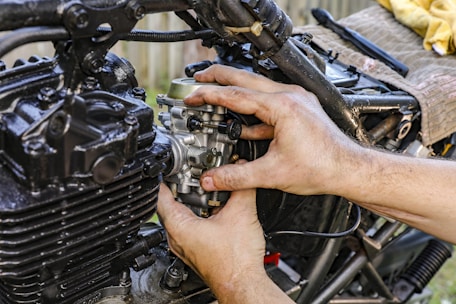 A person’s hands are working on a motorcycle engine, focusing on adjusting or installing a component. The engine appears well-used, with various metallic and dark parts visible, surrounded by bolts, wires, and tubes.