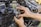 A person’s hands are working on a motorcycle engine, focusing on adjusting or installing a component. The engine appears well-used, with various metallic and dark parts visible, surrounded by bolts, wires, and tubes.