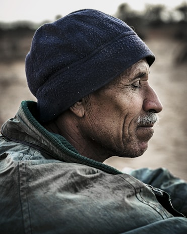 Profile of an older man wearing a dark blue hat and greenish jacket. His face is weathered and he has a contemplative expression.