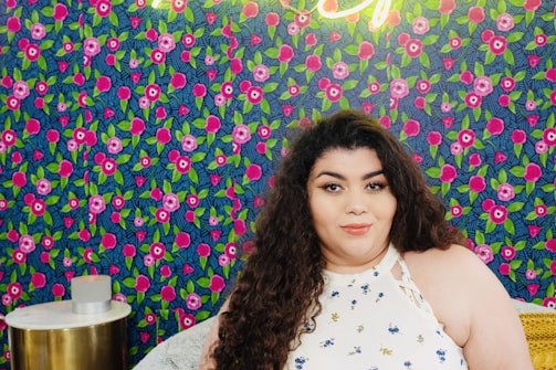 A person with long curly hair wearing a white top with small floral designs is seated in front of a vibrant, colorful wall with a floral pattern featuring pink and green hues. A small round table with a gold base is nearby.