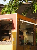 A colorful food truck partially shaded by the surrounding greenery. Inside the truck, a person is visible, wearing a striped shirt. Various objects like bottles and containers are seen on the shelves, and there are illuminated images of food displayed on the truck.