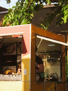 A colorful food truck partially shaded by the surrounding greenery. Inside the truck, a person is visible, wearing a striped shirt. Various objects like bottles and containers are seen on the shelves, and there are illuminated images of food displayed on the truck.