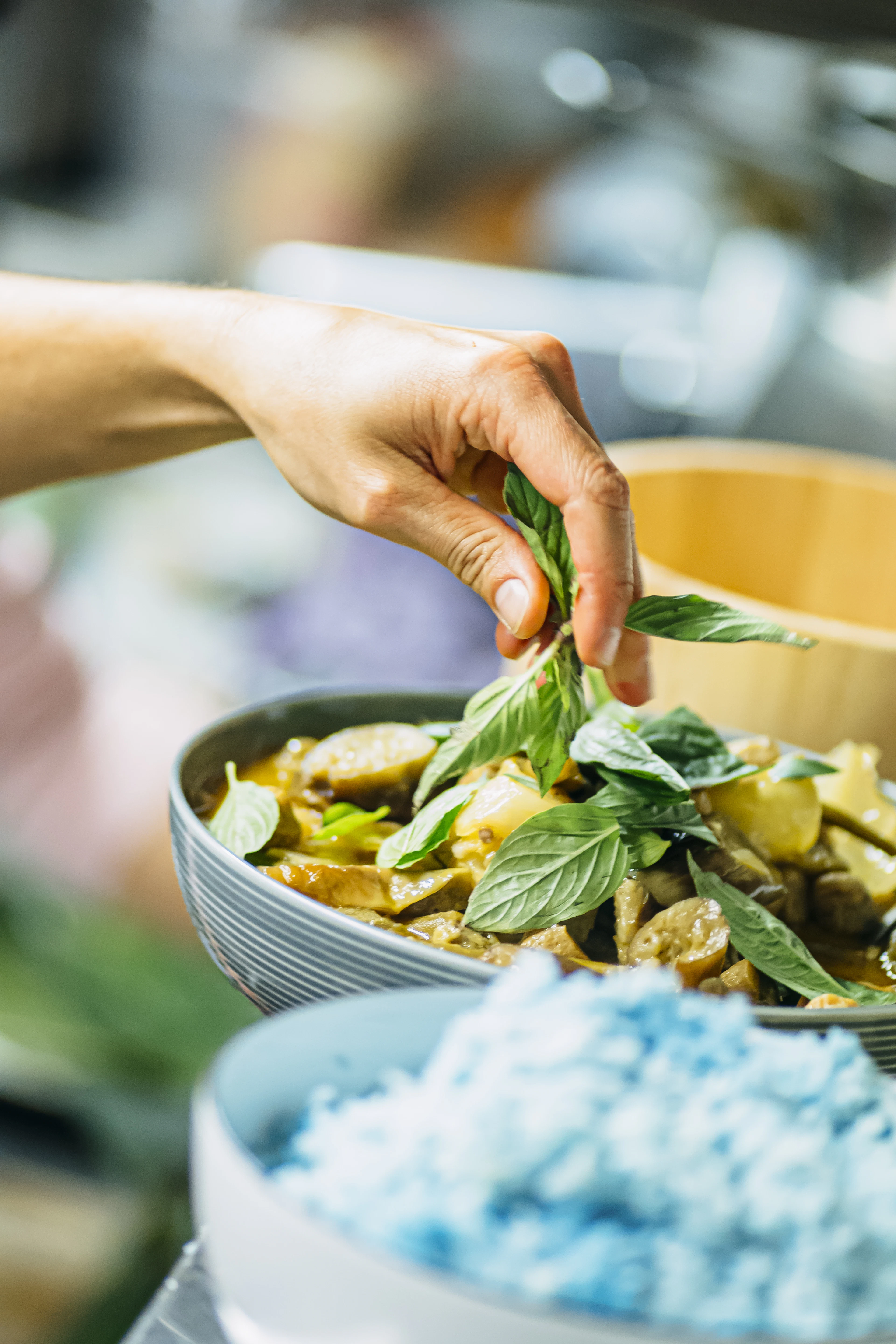 A hand scattering Thai basil over a bowl of green curry with Thai eggplant