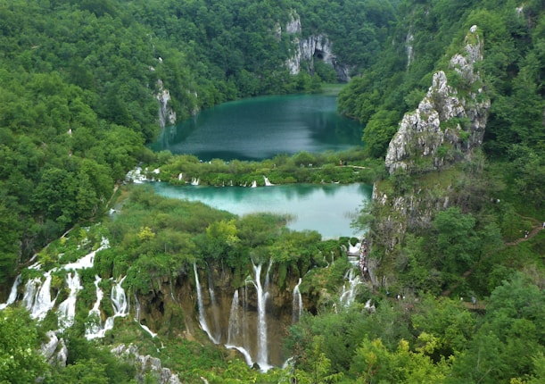 Lush greenery surrounds a series of cascading waterfalls that flow into a serene, turquoise lake nestled in a valley. The landscape features rugged rock formations and dense forests.