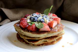 A stack of fluffy protein pancakes garnished with maple syrup and a sprig of mint on a wooden table.