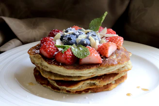 A stack of golden-brown fitness pancakes drizzled with sugar-free syrup and garnished with sliced strawberries on a pastel pink plate.