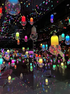 A room filled with colorful, hanging light bulbs reflected in mirrors, creating a kaleidoscopic effect. The bulbs vary in colors such as red, blue, green, yellow, and pink, and the reflections multiply their presence, creating an immersive visual experience. The floor and walls contribute to the reflective ambiance, enhancing the vibrant and playful atmosphere.