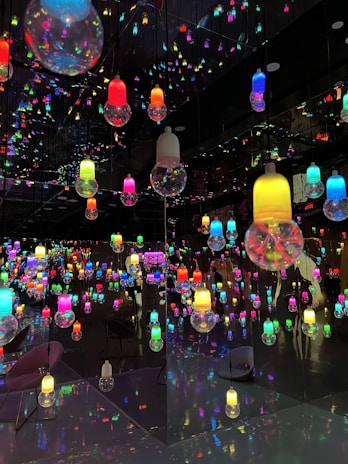 A room filled with colorful, hanging light bulbs reflected in mirrors, creating a kaleidoscopic effect. The bulbs vary in colors such as red, blue, green, yellow, and pink, and the reflections multiply their presence, creating an immersive visual experience. The floor and walls contribute to the reflective ambiance, enhancing the vibrant and playful atmosphere.