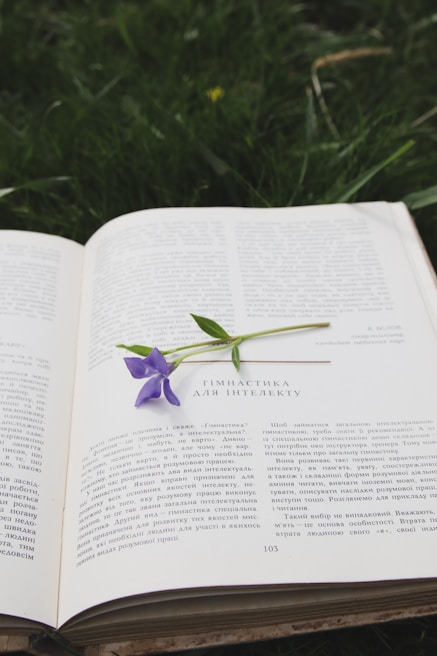 An open book lies on the grass with a purple flower placed on top of it. The text is written in a Cyrillic script, suggesting possible Eastern European content. The setting appears natural and serene with lush green grass surrounding the book.