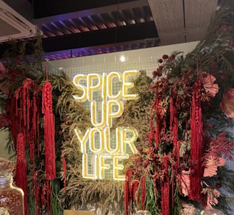A neon sign with the words 'SPICE UP YOUR LIFE' is surrounded by lush greenery and vibrant red flowers and plants. The setting appears to be indoors with a ceiling featuring metallic structures and lighting. There are glass jars in the foreground.
