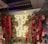 A neon sign with the words 'SPICE UP YOUR LIFE' is surrounded by lush greenery and vibrant red flowers and plants. The setting appears to be indoors with a ceiling featuring metallic structures and lighting. There are glass jars in the foreground.