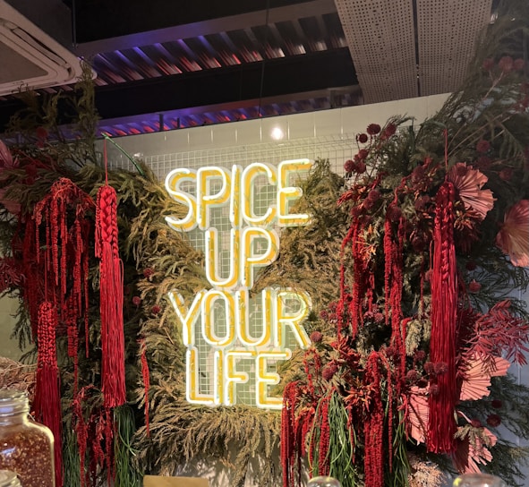 A neon sign with the words 'SPICE UP YOUR LIFE' is surrounded by lush greenery and vibrant red flowers and plants. The setting appears to be indoors with a ceiling featuring metallic structures and lighting. There are glass jars in the foreground.