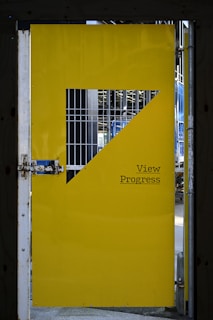 A yellow metal door with a geometric cutout and a phrase 'View Progress' printed on it. The door is partially open, revealing a metal grid and the framework of a construction site or industrial structure behind it. The metal lock and hinges are visible on the side.