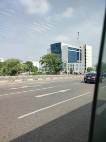 A multi-story modern office building with reflective glass windows stands prominently beside a wide road. The structure is surrounded by lush green trees and bushes, with a partly cloudy sky above. In the foreground, a black car drives along the road, and the street appears calm with minimal traffic.