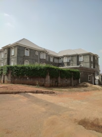 A large, multi-story residential building with a gray exterior and numerous windows featuring arched designs. The roof is light-colored, and the building is surrounded by a high wall covered with dense greenery. The ground in front shows a reddish-brown sandy area with some construction marks.