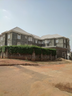 A large, multi-story residential building with a gray exterior and numerous windows featuring arched designs. The roof is light-colored, and the building is surrounded by a high wall covered with dense greenery. The ground in front shows a reddish-brown sandy area with some construction marks.