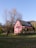 A bright pink house with a gabled roof is situated in an open grassy area. It has solar panels installed on the roof, indicating an eco-friendly design. A tall, leafless tree stands nearby, adding to the rural feel. The background includes a grassy field and a partially visible hill with sparse vegetation, under a clear blue sky.
