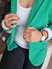 A person wearing a green blazer over a white top is adjusting the blazer with their hands. They have a watch on their left wrist and various rings on their fingers. The person's nails have a light-colored polish, and they are sitting on a piece of furniture.