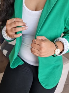 A person wearing a green blazer over a white top is adjusting the blazer with their hands. They have a watch on their left wrist and various rings on their fingers. The person's nails have a light-colored polish, and they are sitting on a piece of furniture.
